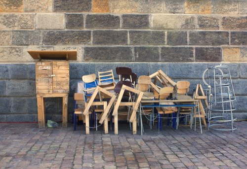 A collection of assorted wooden chairs and small tables piled together and leaning against a grey brick wall, with some chairs stacked in multiple layers and others scattered. The chairs are primarily made of light natural wood, with a few painted or stained darker. To the left, there is a tall, narrow wooden cabinet or enclosure with a lid partially open, situated directly on a cobblestone ground. On the right side, a metal folding chair and a metal wire frame structure are visible, also resting against the brick wall. The scene appears to depict discarded or surplus furniture placed outdoors, possibly as part of a rubbish clearance in Bethnal Green, with the setting illuminated by natural daylight.