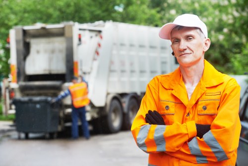 A professional waste management worker wearing an orange high-visibility jacket with reflective silver and orange stripes, stands with arms crossed in front of a large, white skip loader truck. The worker has a neutral expression and a white cap, with a background of green trees and outdoor environment typical of a suburban or urban garden clearance setting. The skip, partially visible, is positioned on a paved or driveway area, with the back panel open, revealing an empty container used for loading garden waste or household rubbish. Behind the worker, another staff member, dressed similarly in an orange high-visibility vest, is seen loading or unloading black rubbish bins into the truck. The scene suggests an organized rubbish removal operation, typical of gardens or property clearance services offered by Garden Clearance Bethnal Green, serving the Bethnal Green area and nearby postcode regions. The environment appears bright and clear, emphasizing a professional and efficient waste collection process, aligned with local rubbish clearance services.
