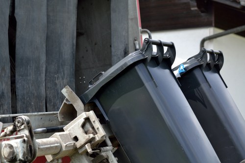 Close-up of two black plastic rubbish bins with blue-lidded openings, mounted on a metal frame attached to a collection vehicle, likely used for waste collection in Bethnal Green. The bins are positioned in the foreground, showing textured, matte surfaces with slight reflections of ambient light. Behind the bins, part of the vehicle's metallic structure and a wooden panel are visible, indicating a street or driveway environment typical of urban waste management sites. The setting suggests routine rubbish collection services provided by Garden Clearance Bethnal Green, supporting local waste disposal needs in the area. The image captures the practical aspect of rubbish removal with a focus on the bin design and their placement for collection, emphasizing the company's role in maintaining cleanliness in the community.