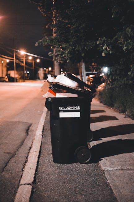 A black wheeled rubbish bin positioned on a sidewalk next to a curb on a quiet street at night. The bin is branded with the name 'St. John's' and has an open lid revealing various discarded items, inc